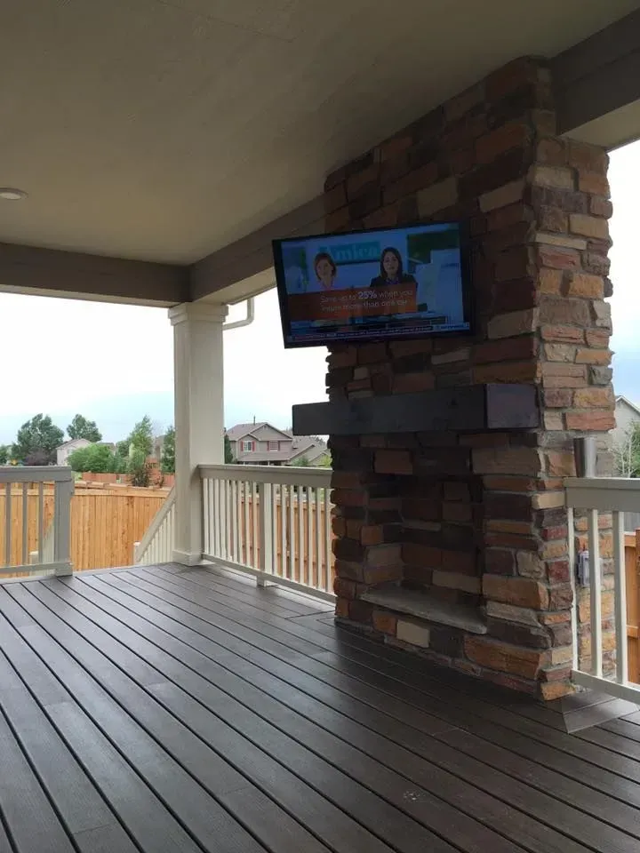 Outdoor deck with a mounted TV on a stone pillar; wooden deck, white railing, houses in the background.