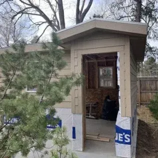 Beige shed with an open doorway, blue wrap, and a tree in front.