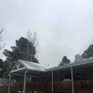 A snow-covered house with a porch and trees against a gray, overcast sky.