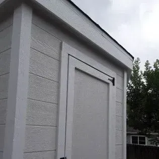 Gray shed with white trim and a black handle against a cloudy sky.