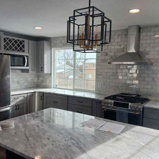 Modern kitchen with grey cabinets, marble countertops, stainless steel appliances, and a geometric chandelier.