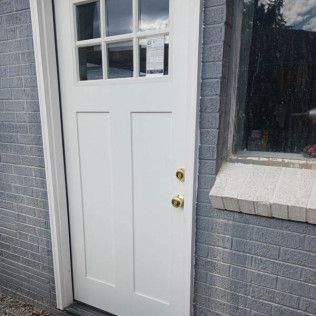 White door with glass pane top and brass door handle, next to a window on a gray brick building.
