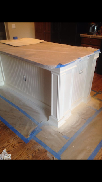 White kitchen island being painted, covered in painter's tape and protective sheeting, set on a hardwood floor.