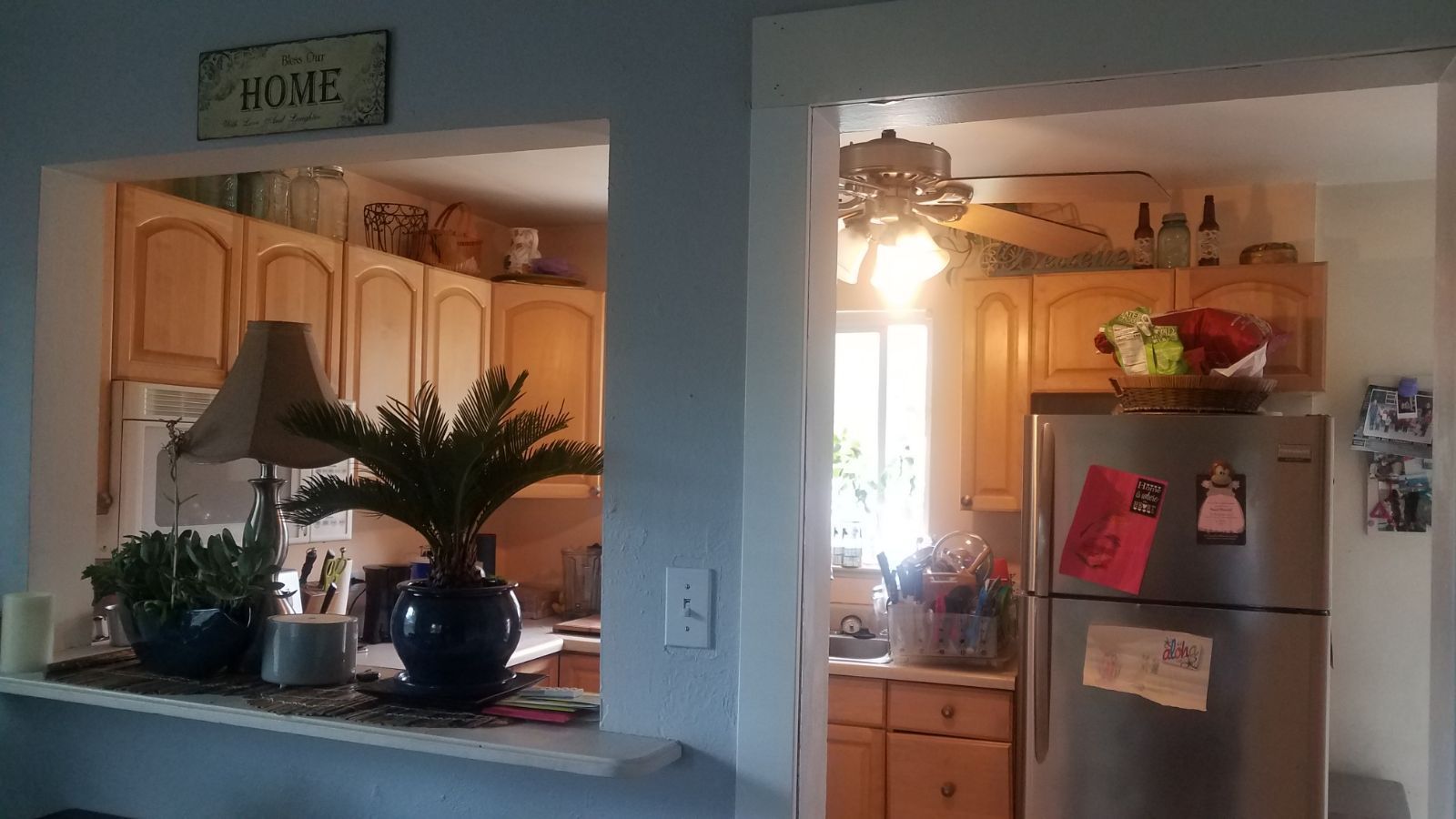 View into a kitchen with light wood cabinets, open pass-through. Plants and lamp on a ledge.