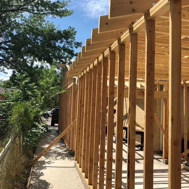 Wooden framework of a building under construction, exterior view with a sidewalk and foliage.