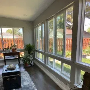 Sunroom interior with a row of windows overlooking a fenced yard. Dark wood floors, plants, and seating are visible.