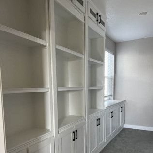 White built-in bookshelves with cabinets, next to a window with blinds, in a room with gray walls and carpet.