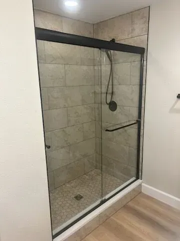 A walk-in shower with beige tile walls and a sliding glass door. A dark-colored showerhead and handle are visible.