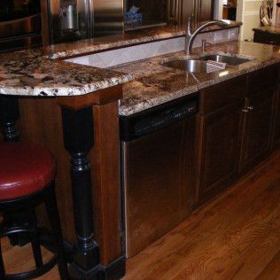 Kitchen island with granite countertop, sink, dishwasher, dark cabinets, and bar stool.