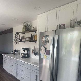 Kitchen with white and gray cabinets, stainless steel refrigerator, and open shelving with plants and jars.