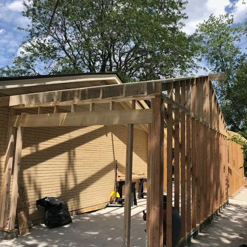 Wooden frame construction of a building extension, beige siding, blue sky, trees.
