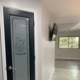 Pantry door with frosted glass and pantry label. Black frame with silver doorknob. Adjacent wall with TV and window.