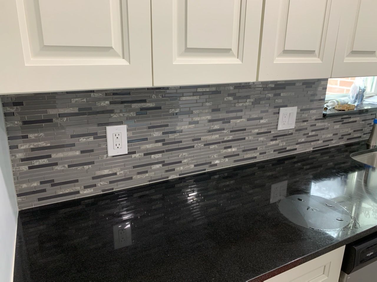 Kitchen with black countertop, white cabinets, and gray tiled backsplash. Two electrical outlets are visible.