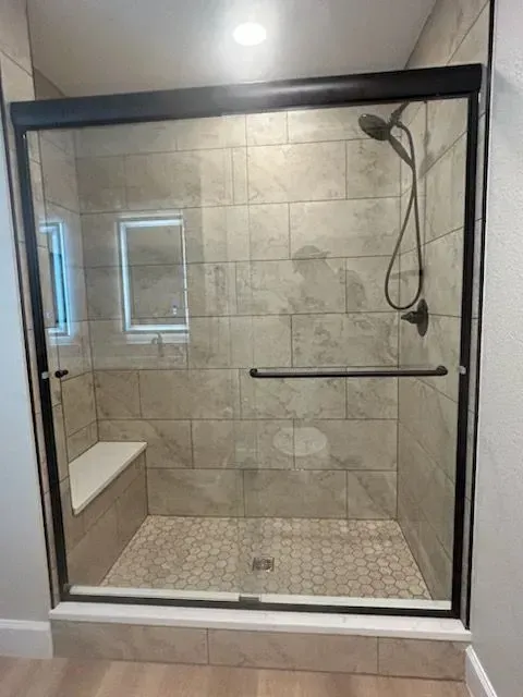 Shower stall with glass door, black frame, gray tile, showerhead, and a built-in bench.
