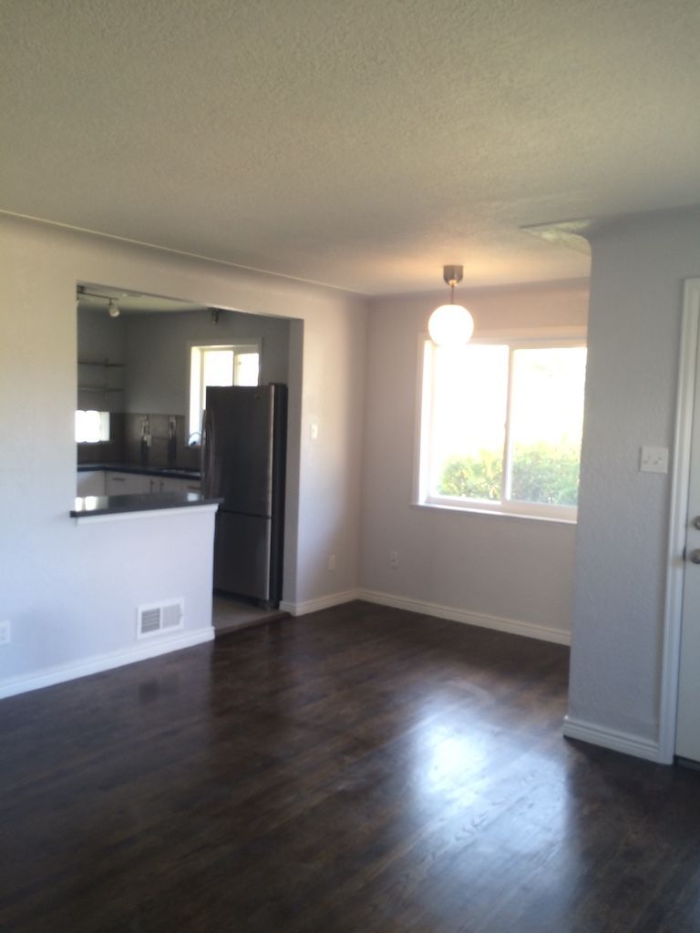 An empty dining room with dark wooden floors, gray walls, and a view into a kitchen with a refrigerator.