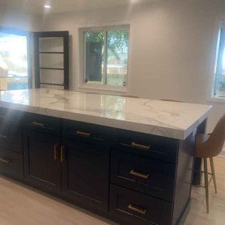 Dark kitchen island with marble countertop, gold hardware, and a brown stool in front of a window.