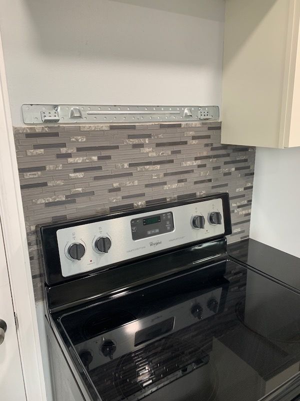 A kitchen with a Whirlpool electric stove, backsplash, and upper cabinets; a silver metal mounting bracket is on the wall.