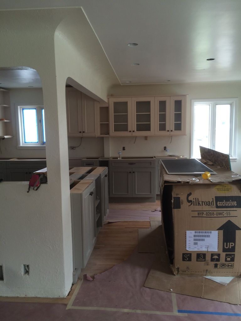Kitchen renovation in progress: cabinets installed, appliance box, light beige walls, wooden floors, windows.