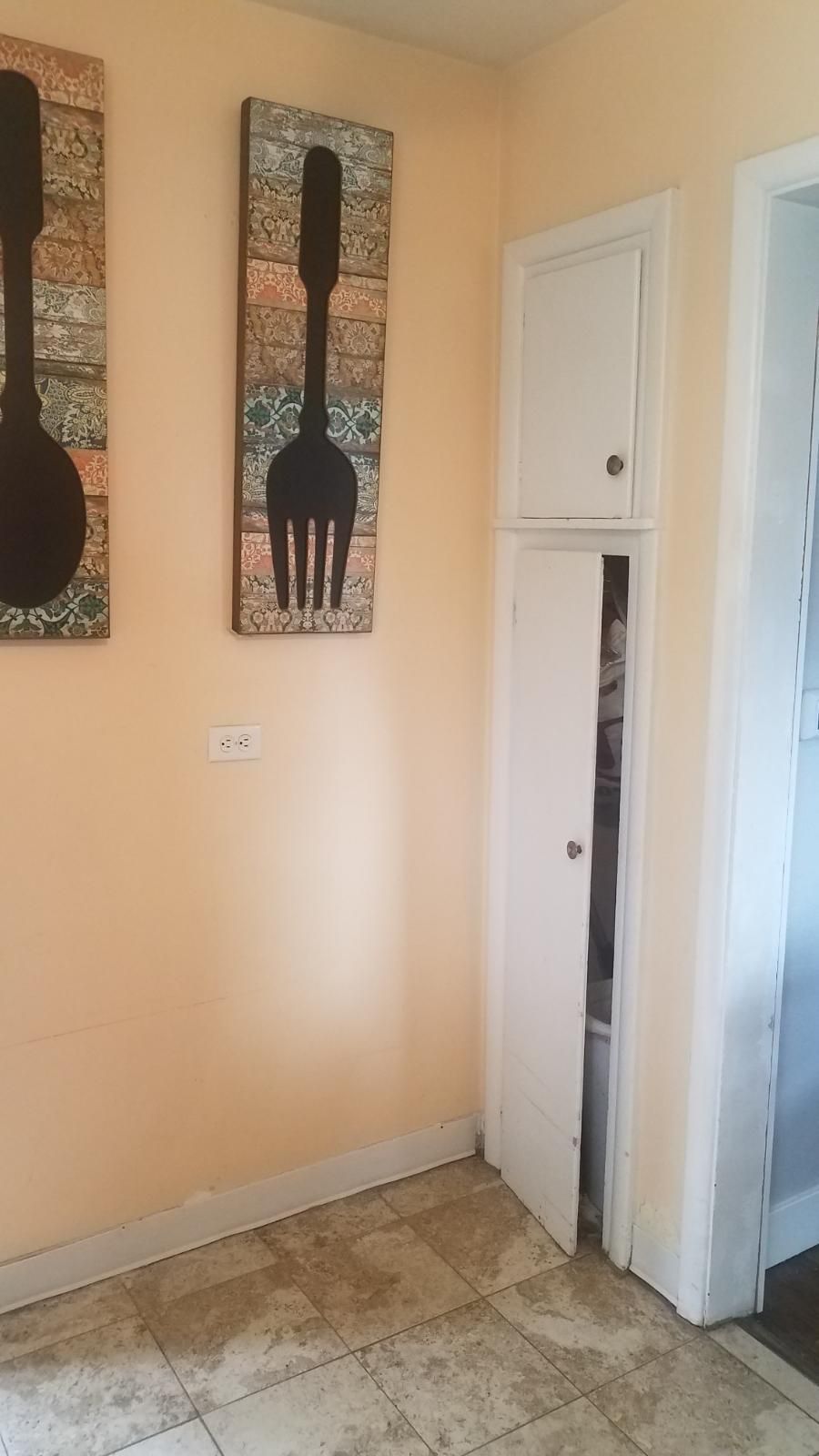 Narrow white pantry with a partially open door, next to a doorway and two decorative utensil art pieces.