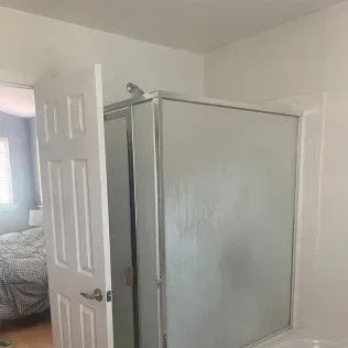 White door open to a bathroom with a shower stall. Bedroom visible in the background.