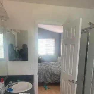 Bathroom interior, showing a sink, mirror, and doorway leading to a bedroom with a bed.