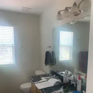 Bathroom with toilet, sink, mirror, blinds, towels, and toiletries.