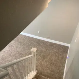 View from top of stairs. White railing leads down to carpeted landing with gray walls.