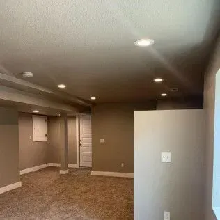 Basement interior with recessed lighting, tan walls, carpet, and a white door.