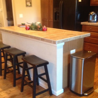 Kitchen island with tiled countertop, stools, and stainless steel trash can.