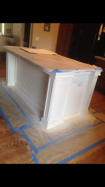 White kitchen island being painted, covered in blue painter's tape and plastic sheeting on a hardwood floor.