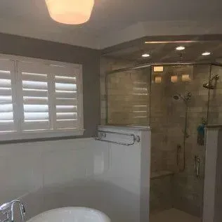 Bathroom with white tub, shower with glass doors, and window with shutters.