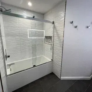 White-tiled bathroom with a glass shower door over a bathtub. Dark floor, white walls, and silver fixtures.