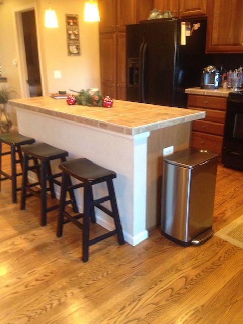 Kitchen with island, four bar stools, stainless steel trash can, hardwood floors, and black refrigerator.