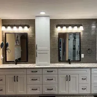 Bathroom with dual sinks, mirrors, and a tall cabinet. Gray tiled wall, white cabinets, and countertop.