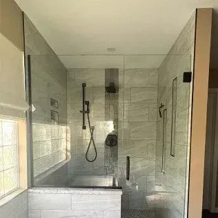 Modern glass shower enclosure with gray tile walls, dark fixtures, and window.