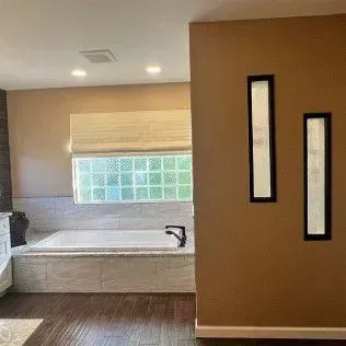 Bathroom with a tan wall, bathtub, window with frosted glass, and two tall, framed art pieces.