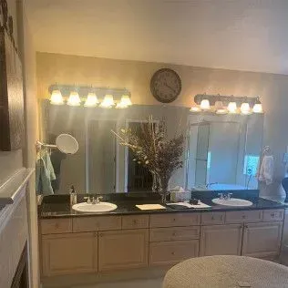 Bathroom with dual sinks, mirrors, and beige cabinets; a vase of dried flowers and a round clock are also present.