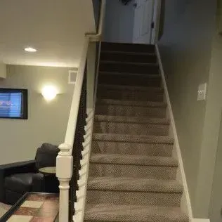 Staircase with carpeted steps leading upward; handrail on the left, dark chair and lamp at the bottom.