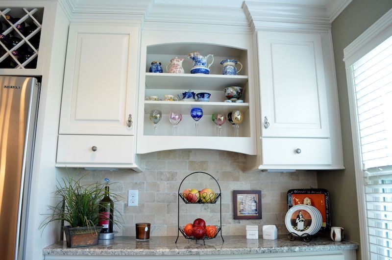 Kitchen cabinets with open shelves displaying dishware and stemware above a countertop with fruit and decor.