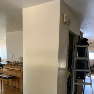 Cream-colored column in a room. A ladder leans against a doorway. A piano is visible in the background.