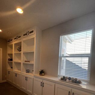 Built-in white bookshelves with cabinets, next to a window with blinds, in a room.