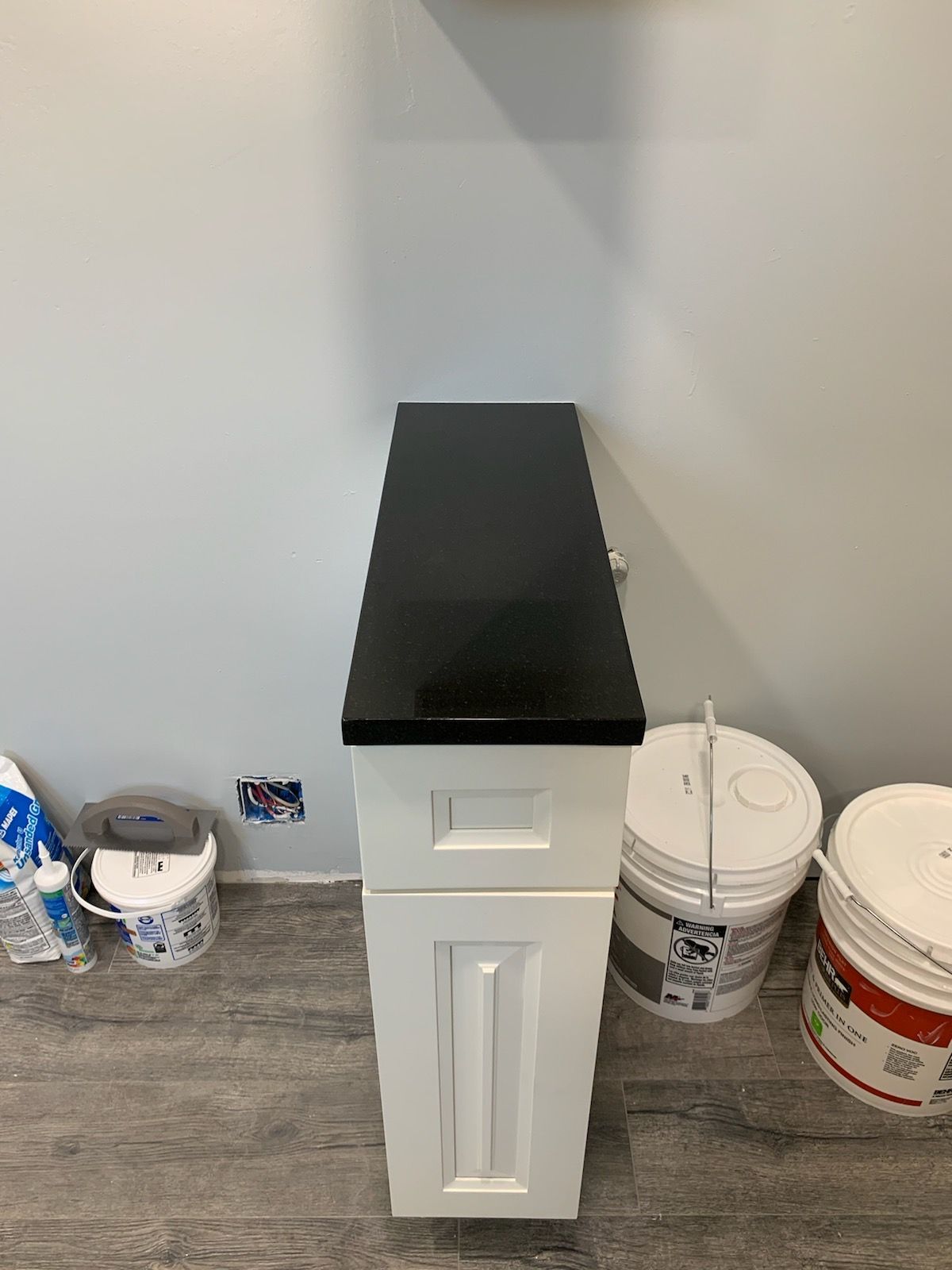 White cabinet with black countertop against a gray wall. Paint buckets and supplies are on the floor.