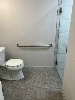 Bathroom with toilet, grab bar, and glass shower door; gray tile floor and white walls.