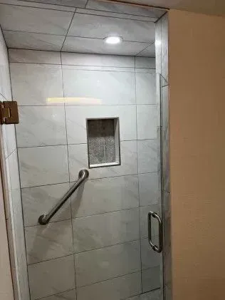 Shower stall with glass door, white tiled walls, grab bar, and recessed shelf.