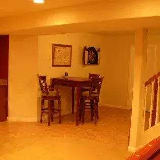 Basement room with a small table and chairs, dartboard, and stairs. Warm lighting.
