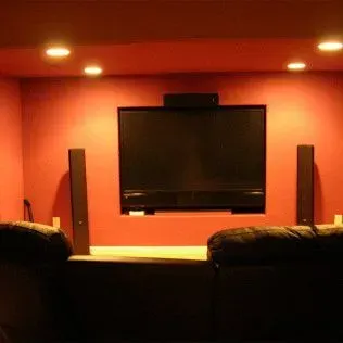 Television mounted on red wall with two speakers, recessed lighting and seating.