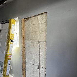 Opening in gray wall reveals concrete block wall; new door leaning against left wall.