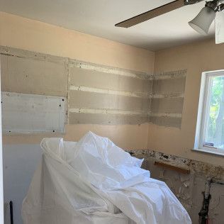 Bathroom under renovation with exposed drywall and covered fixtures, tan walls, and a window.