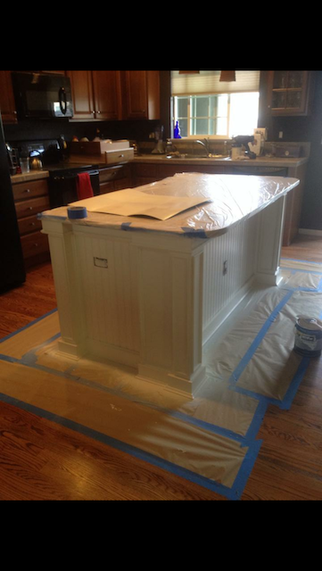 Kitchen island being painted, covered in protective plastic and painter's tape; wood floor.