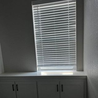 Window with closed white blinds above a white cabinet. Sunlight streams through the blinds.